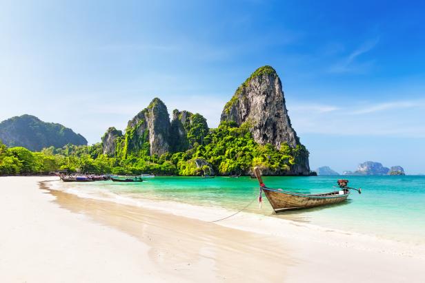 Ein traditionelles thailändisches Boot liegt an einem tropischen Strand mit Kalksteinfelsen im Hintergrund.