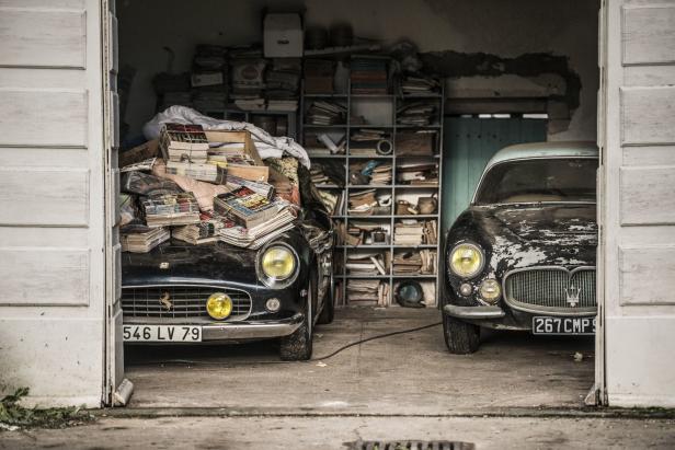 In einer Garage stehen zwei Oldtimer, einer davon mit einem Stapel Zeitschriften bedeckt.