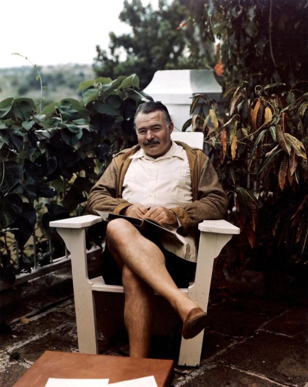 Ernest Hemingway sitzt mit einem Buch in einem weißen Stuhl im Freien.