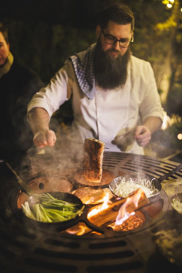 Ein Koch mit Bart grillt Fleisch und Gemüse auf einem offenen Feuer.