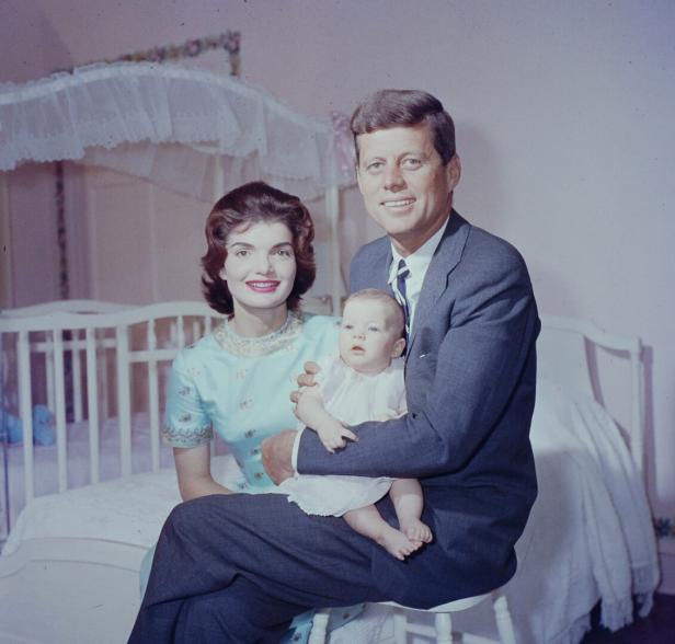 John F. Kennedy, Jackie Kennedy und ihr Baby posieren vor einem Kinderbett.