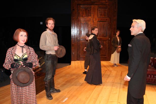 Szene aus einem Theaterstück mit mehreren Darstellern in historisch anmutenden Kostümen auf einer Holzbühne.
