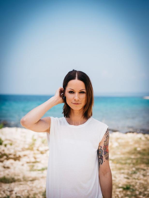 Christina Stürmer vor einem Strand mit blauem Meer im Hintergrund.