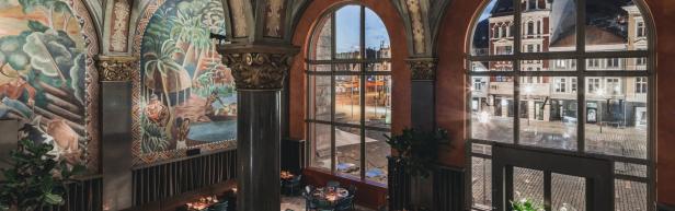 Innenansicht eines Restaurants mit Wandmalereien und Blick durch große Fenster auf eine belebte Straße.