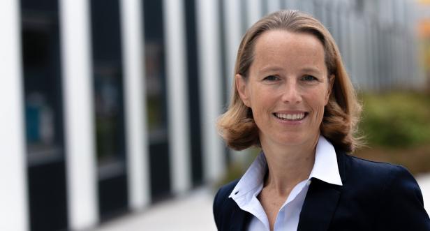 Eine lächelnde Frau in einem Blazer vor einem modernen Gebäude.