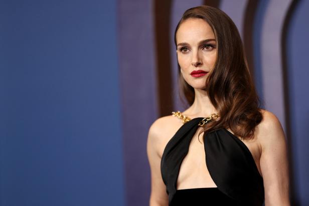Natalie Portman in einem schwarzen Kleid mit goldenen Ketten vor einem blauen Hintergrund.