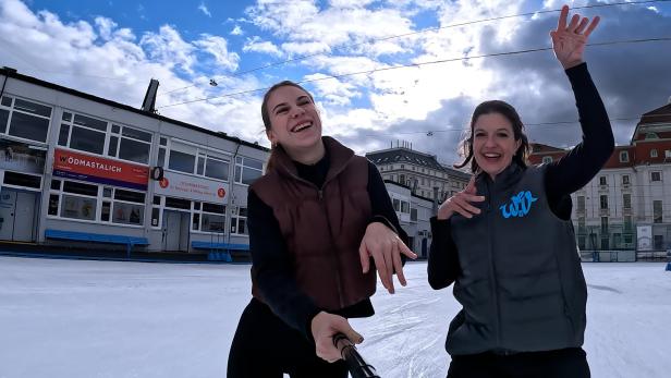 Zwei Frauen machen ein Selfie auf einer Eisbahn vor einem Gebäude.