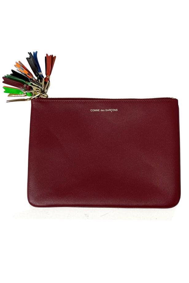 Eine bordeauxrote Clutch von Comme des Garçons mit einem bunten Quastenanhänger.