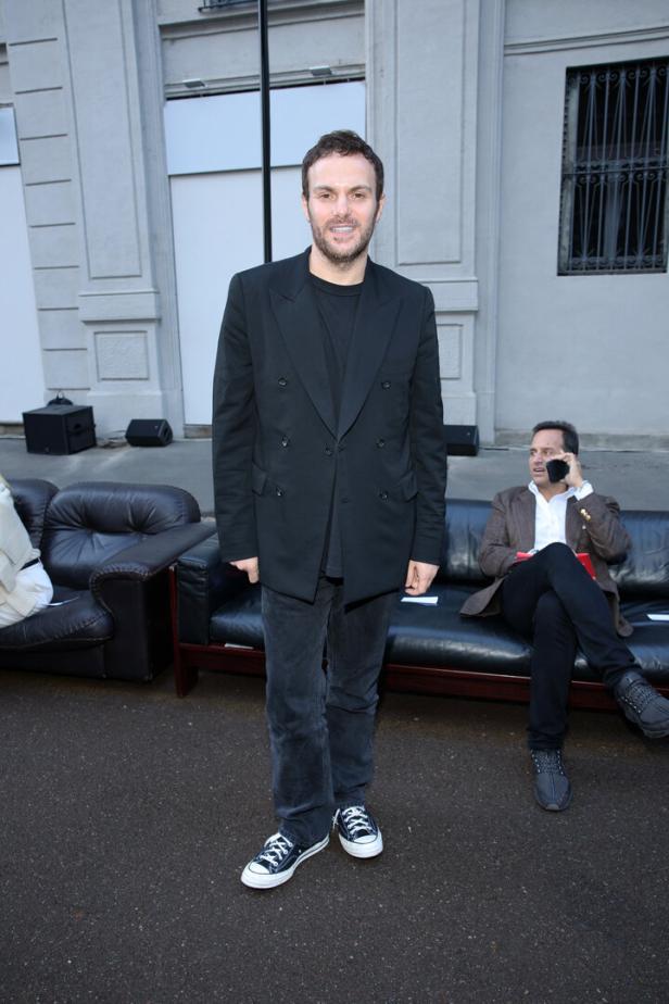 Ein Mann in einem schwarzen Anzug und Converse-Turnschuhen steht vor einem Gebäude.