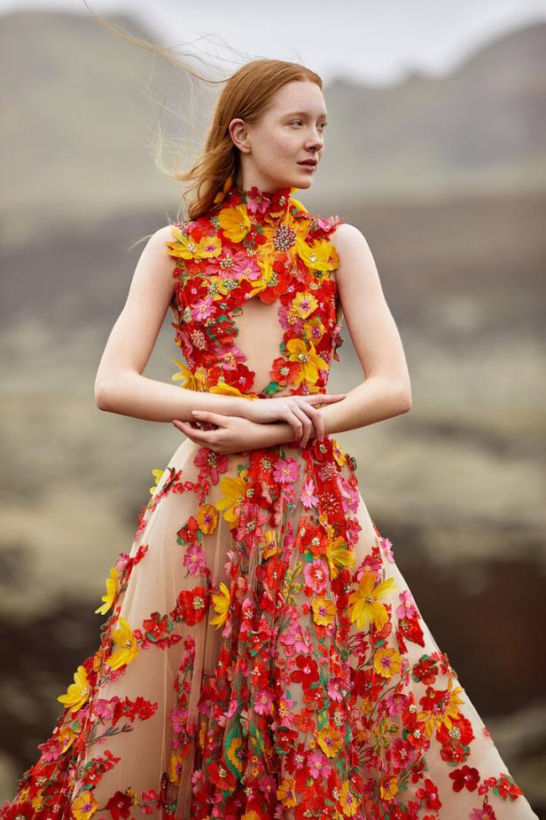 Eine Frau mit roten Haaren trägt ein langes Kleid, das mit roten und gelben Blumen verziert ist.