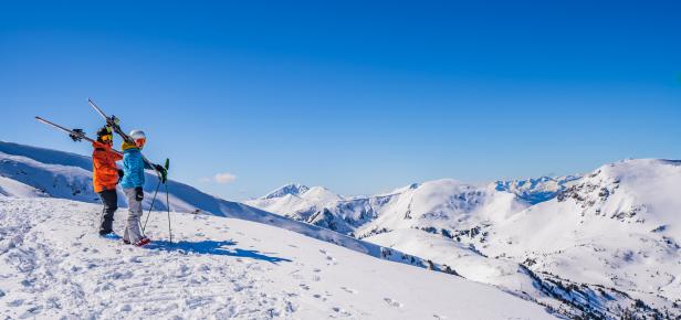 Zwei Skifahrer stehen auf einem schneebedeckten Berg und blicken auf die Gipfel.