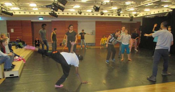 Eine Gruppe von Menschen probt auf einer Bühne mit schwarzem Boden.