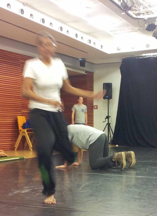 Eine Frau tanzt barfuß, während andere Personen im Hintergrund zu sehen sind.