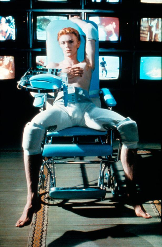 David Bowie sitzt in einem blauen Stuhl, umgeben von Fernsehbildschirmen.