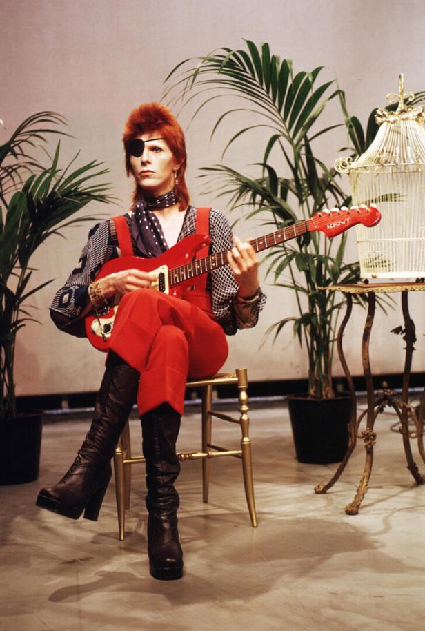 David Bowie mit Augenklappe, rotem Overall und Gitarre, sitzt auf einem Stuhl.