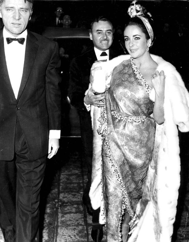 Elizabeth Taylor in einem gemusterten Kleid und einer Stola mit zwei Männern.