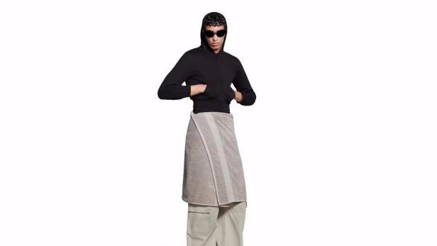 Eine Person mit Sonnenbrille, Kapuzenpullover und einem um die Hüften gewickelten Handtuch steht vor einem weißen Hintergrund.