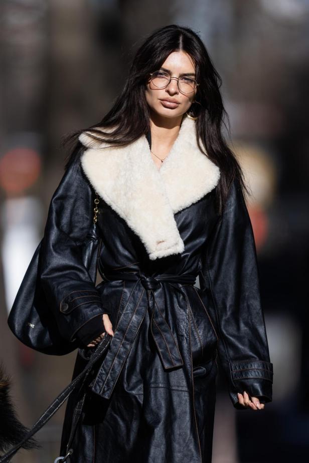Emily Ratajkowski mit schwarzem Mantel in Lederoptik und großer, runder Brille.