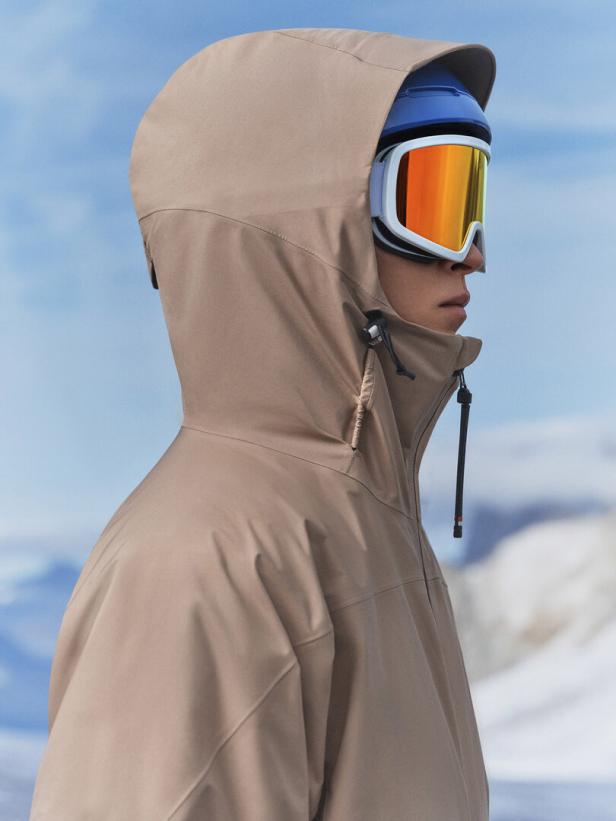 Eine Person mit Skibrille und Kapuze vor einem verschneiten Hintergrund.