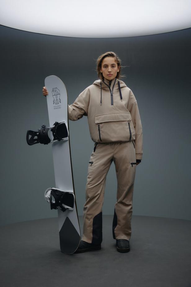 Eine Frau in beiger Skibekleidung posiert mit einem Snowboard.