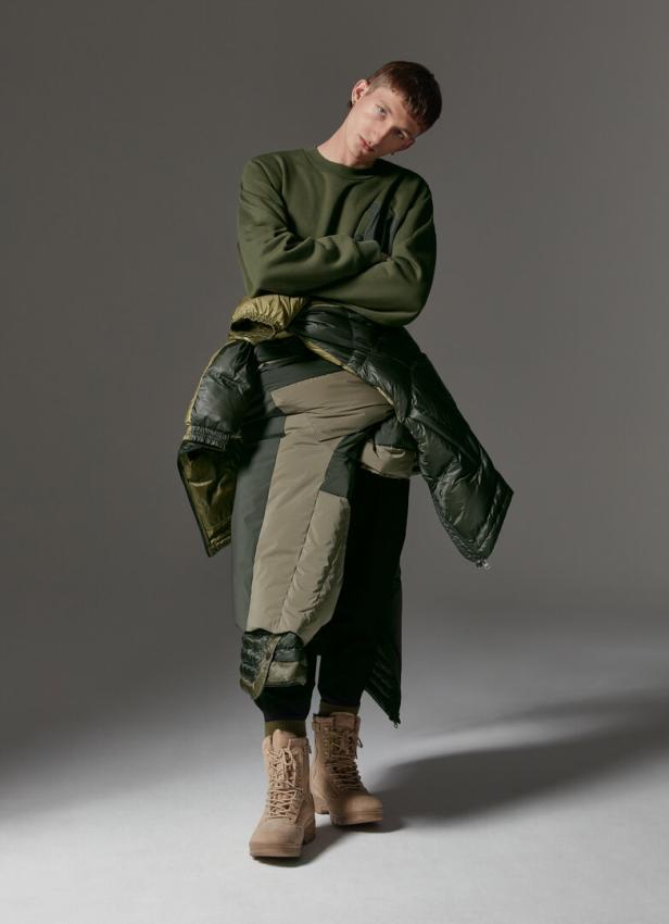 Ein Mann mit verschränkten Armen trägt einen grünen Pullover und eine Jacke über der Schulter, dazu Stiefel.
