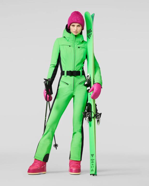 Eine Frau in einem grünen Skianzug mit pinkfarbener Mütze, Handschuhen und Stiefeln hält grüne Ski.