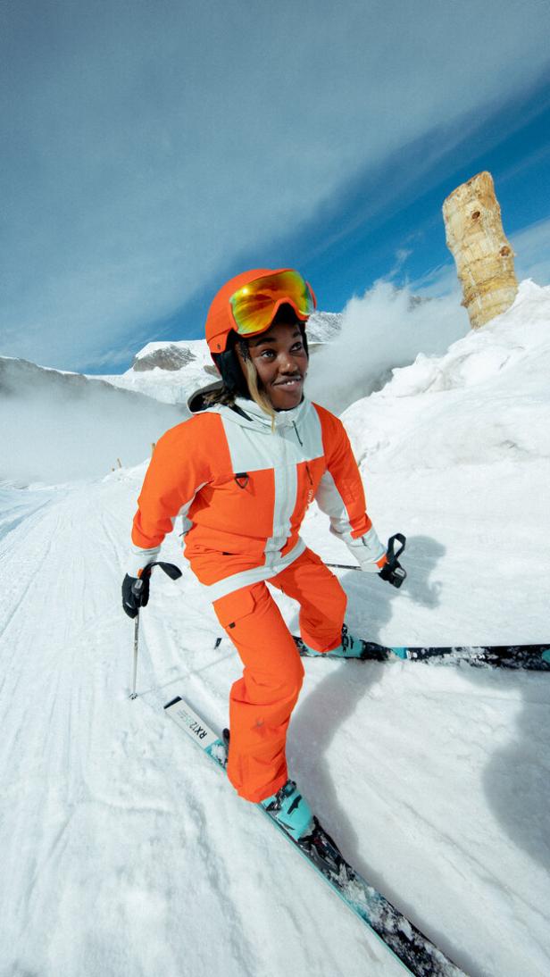 Eine Person in orangefarbener Skikleidung fährt Ski auf einer schneebedeckten Piste.