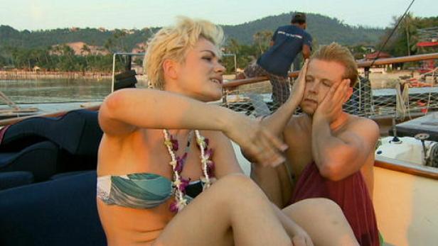 Eine Frau mit Blumenkette berührt das Gesicht eines Mannes auf einem Boot.