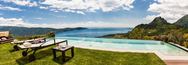 Ein Infinity-Pool mit Blick auf einen See und grüne Hügel an einem sonnigen Tag.