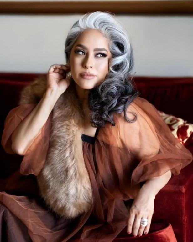 Claudia Fabian posiert mit grauem Haar und einer Pelzstola auf einem roten Sofa.