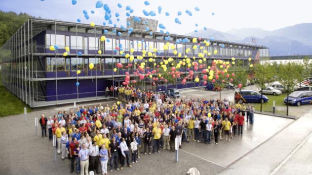 Eine große Gruppe von Menschen steht vor einem modernen Bürogebäude, umgeben von bunten Luftballons.
