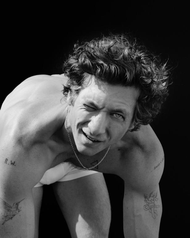 Ein Mann mit lockigem Haar und Tattoos zwinkert in die Kamera.