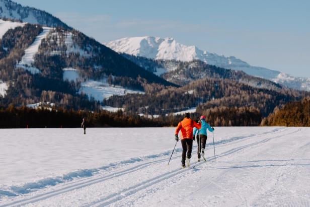 Zwei Personen laufen auf Skiern durch eine verschneite Landschaft mit Bergen im Hintergrund.
