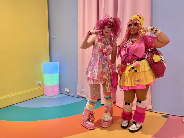 Zwei Frauen in farbenfrohen, verspielten Outfits posieren vor einem bunten Hintergrund.