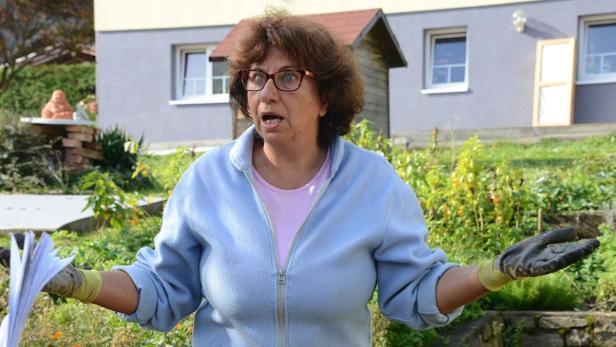Eine Frau mit Brille gestikuliert in einem Garten mit einem Stapel Papier in der Hand.