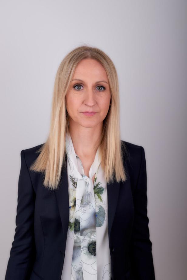 Eine Frau mit blonden Haaren trägt ein dunkles Jackett und eine Bluse mit Blumenmuster.