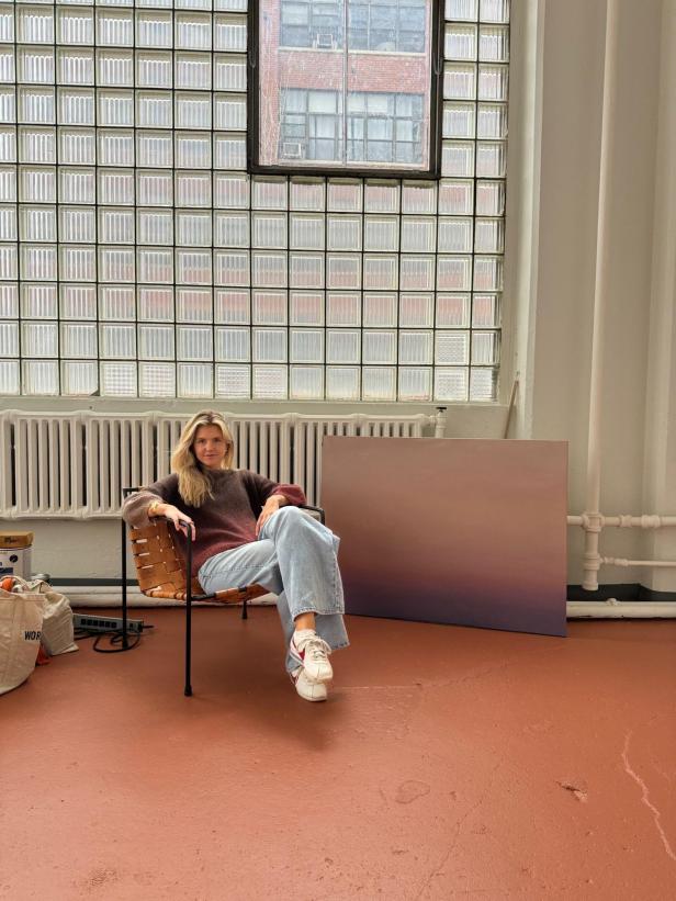 Eine blonde Frau sitzt auf einem Stuhl. Hinter ihr sieht man ein Kunstwerk, das vor einem Heizkörper und einer Fensterscheibe angelehnt ist