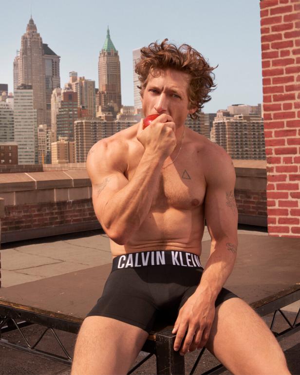 Ein Mann mit Calvin Klein Unterwäsche sitzt auf einem Dach und isst einen Apfel vor der Skyline von New York.