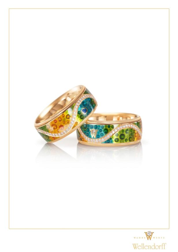 Zwei goldene Wellendorff-Ringe mit farbiger Emaille und Diamanten liegen auf einer weißen Oberfläche.