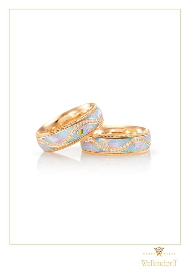 Zwei goldene Wellendorff-Ringe mit pastellfarbenem Emaille und Diamanten liegen auf einer reflektierenden Oberfläche.
