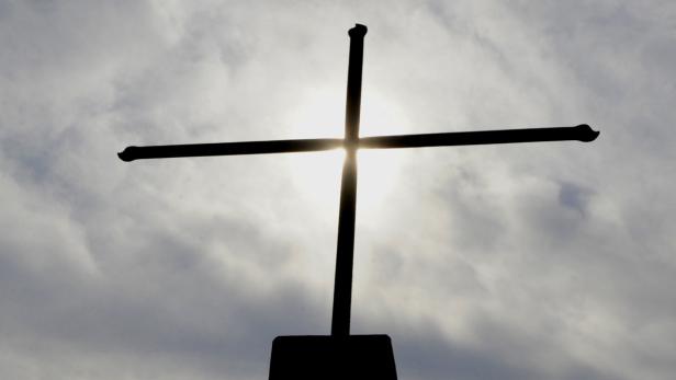 Ein dunkles Kreuz steht vor einem hellen, bewölkten Himmel.