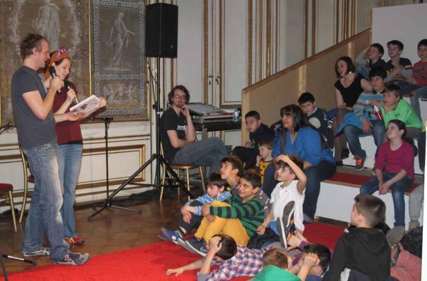 Eine Frau mit Krone und ein Mann lesen vor einer Gruppe von Kindern aus einem Buch vor.