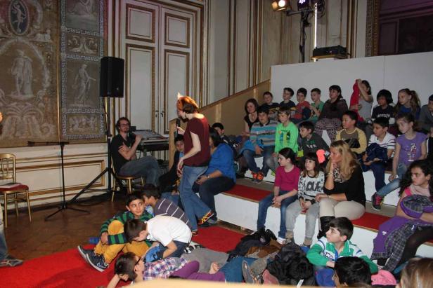 Eine Frau mit Mikrofon spricht vor einer Gruppe von Kindern in einem verzierten Raum.