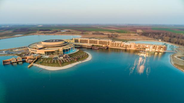 Luftaufnahme eines Resorts mit einem runden Gebäude und einem Hotelkomplex an einem See.