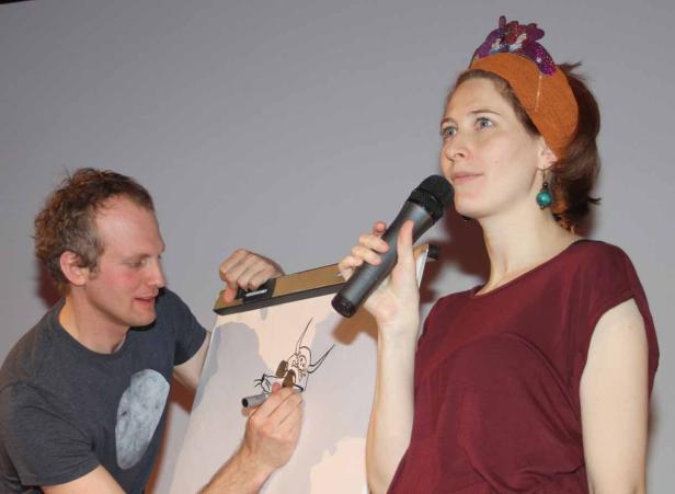 Eine Frau mit Mikrofon steht neben einem Mann, der eine Comicfigur zeichnet.