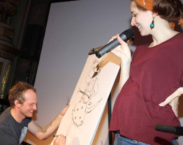 Ein Mann zeichnet eine Karikatur, während eine Frau mit Mikrofon daneben steht.