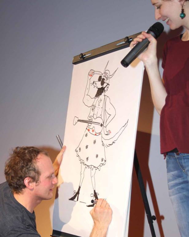 Ein Mann zeichnet eine Karikatur einer Figur mit Hörnern und Rock, während eine Frau daneben ins Mikrofon spricht.