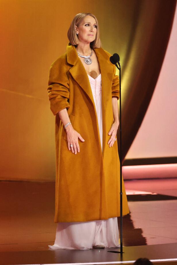 Céline Dion steht auf einer Bühne in einem langen, ockerfarbenen Mantel und einem hellen Kleid.
