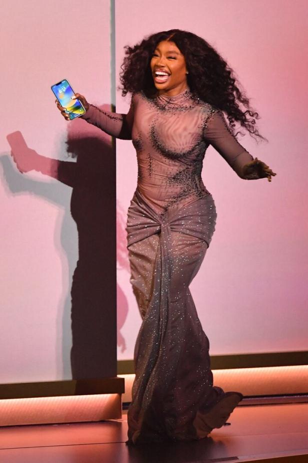 SZA lächelt auf einer Bühne und hält ein Handy in der Hand.