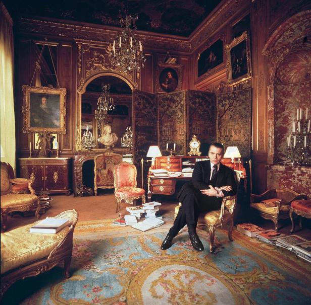 Ein Mann im Anzug sitzt in einem opulenten, mit Kunstwerken gefüllten Raum.